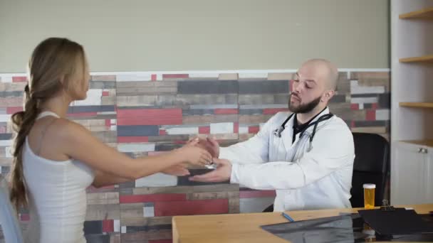 30 años médico barbudo hacer gesto de aliento tomar la mano de una paciente chica en el consultorio médico estrechar la mano y calmarla 4k — Vídeos de Stock