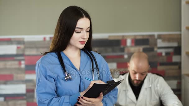 Portrait d'une infirmière aux cheveux bruns avec planche derrière le médecin — Video