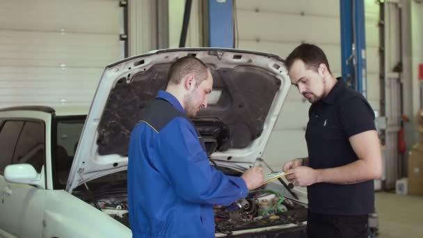 Mecánico del coche está discutiendo con el cliente en el servicio de reparación de automóviles con portapapeles detrás de la campana abierta de un coche — Vídeos de Stock