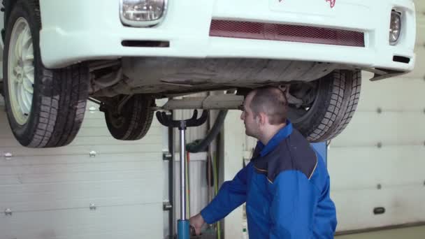 自動車修理工が車のサスペンションを持つ車の修理店で青いオーバー オールにリフトしたクロス オーバー車をチェックします。 — ストック動画