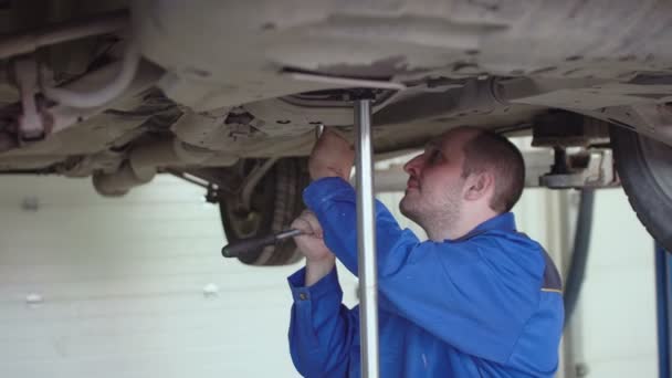 Автомеханик проверяет подвеску на поднятой кроссовер машине в синем комбинезоне в автомастерской. — стоковое видео