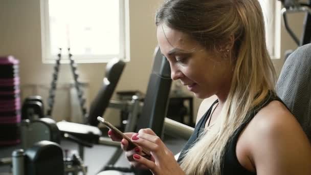 Cansado después del entrenamiento de la belleza de los años 20 mujer deportiva está charlando con el teléfono inteligente en un gimnasio de la máquina — Vídeo de stock