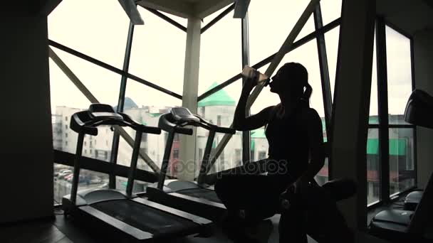 Siluett av kvinna är dricka vatten från sportig flaska i ett gym bakom ett fönster — Stockvideo