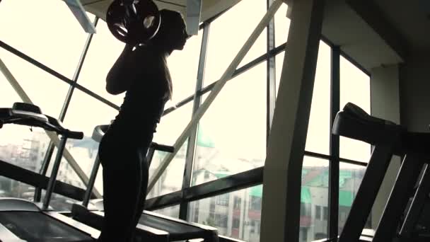 Silueta de una mujer deportiva está levantando la barra detrás de la ventana en un gimnasio — Vídeos de Stock