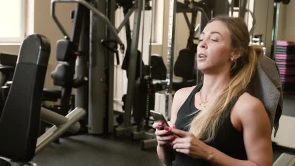 20'li yaşlarda yorgun sarışın sporcumuz çatırdamak ile smartphone bir spor salonunda egzersiz sonrası — Stok video