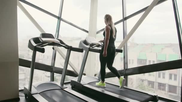 20s beauty sportive woman in black sportswear is running on treadmill and smiling in slow motion behind sunny window — Stock Video