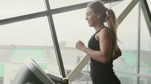 20s belleza deportiva mujer en ropa deportiva negro se ejecuta en la cinta de correr y sonriendo en cámara lenta detrás de la ventana soleada — Vídeo de stock