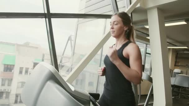 20s beauty sportive woman in black sportswear is running on treadmill and smiling in slow motion behind sunny window — Stock Video