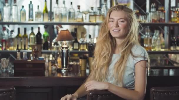 Красивая блондинка в вечернем платье сидит возле барной стойки и смотрит в камеру — стоковое видео