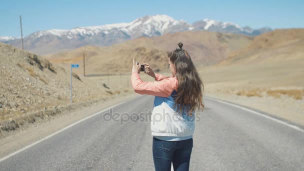 Mujer turista atractiva joven está tomando fotos en la carretera con montañas en el fondo — Vídeos de Stock