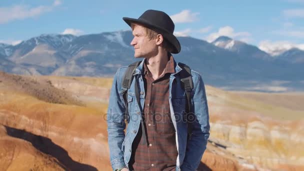 Retrato de viajero hipster en sombrero negro mirando hacia otro lado con montañas en el fondo — Vídeos de Stock