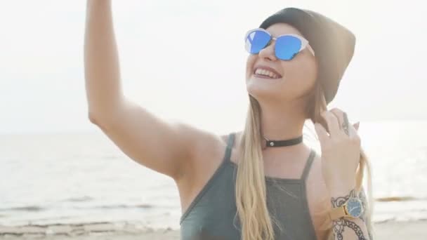 若い金髪サングラスでタトゥーとは海と日没時間のクローズ アップでは、ビーチの背景に電話で selfie. — ストック動画