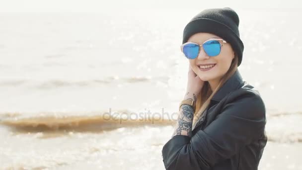 Eine junge Blondine mit Sonnenbrille, schwarzem Hut und Lederjacke, die Wein vor dem Hintergrund von Wasser in Großaufnahme trinkt. — Stockvideo