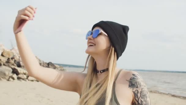 Attraktive tätowierte blonde Mädchen mit Sonnenbrille und schwarzem Hut macht Selfie auf einem Handy am Strand in Großaufnahme. — Stockvideo