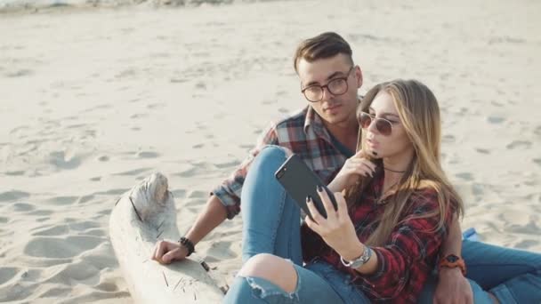 在太阳镜和戴眼镜的家伙的年轻迷人金发女郎坐在一个日志特写镜头上做自拍照在沙滩上. — 图库视频影像