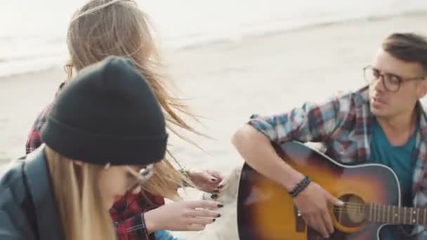 Aantrekkelijke blonde lacht en klapt haar handen, een man met bril speelt gitaar zittend op een logboek op de kust-close-up. — Stockvideo