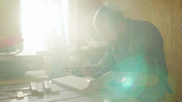 20s бородатий тесляр дерев'яний працівник в фартусі малює на дерев'яній дошці з сонячними спалахами підсвічування — стокове відео