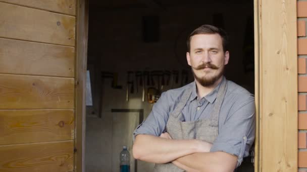 Portrait of 20s bearded woodworker looking at camera, stay behind workshop — Stock Video