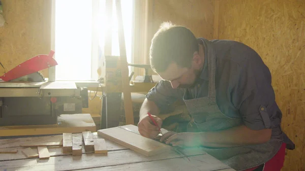 20s barbudo hombre carpintero dibujo sobre tabla de madera. día soleado en el taller —  Fotos de Stock