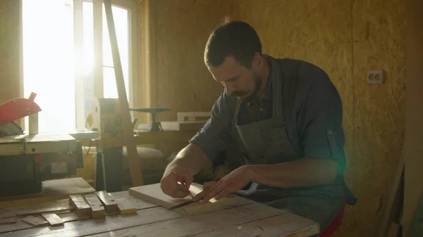 Carpintero barbudo años 20 carpintero carpintero en delantal dibujo sobre tabla de madera con destellos de sol de contraluz — Foto de Stock