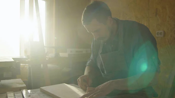 20'li yaşlarda marangoz woodworker arka ışık güneş fişekleri ile ahşap tahta üzerinde çizim önlük sakallı