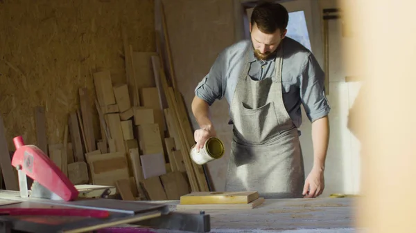 Portrait of 20s bearded carpenter woodworker rubs lacquer in a wooden plank in the workshop Stock Photo