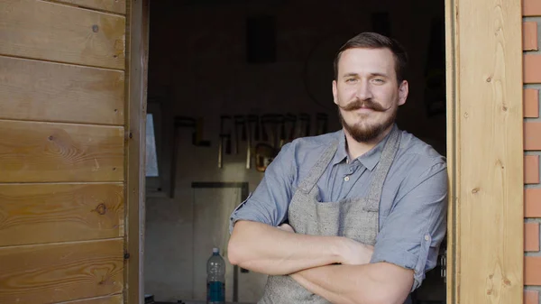 Portrait of 20s bearded woodworker looking at camera, stay behind workshop Stock Photo