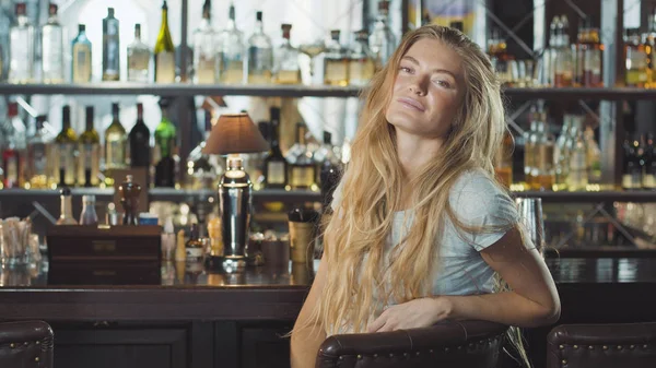 Krásná blondýnka ve večerních šatech sedí u baru a při pohledu na fotoaparát — Stock fotografie