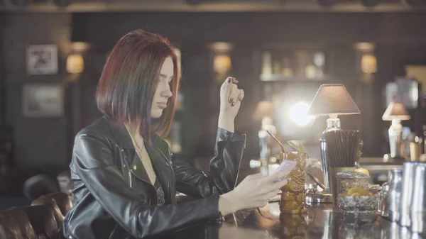 20'li seksi güzellik Kızıl saçlı kadın siyah deri ceketli mi içki kokteyl, kamera ve flört gülümsüyor bardaki bakarak