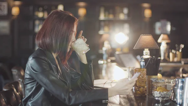 20'li seksi güzellik Kızıl saçlı kadın siyah deri ceketli mi içki kokteyl, kamera ve flört gülümsüyor bardaki bakarak