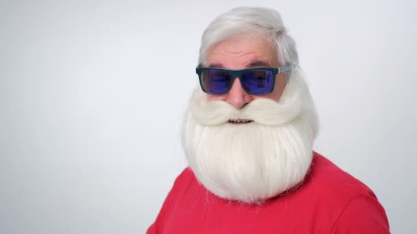Крупним планом красивий хіпстер Санта в сонцезахисних окулярах з великою білою бородою дивиться на камеру на білому тлі — стокове відео