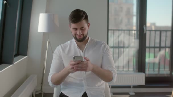 Sonriente joven con camisa blanca está charlando en las redes sociales utilizando el teléfono inteligente — Vídeo de stock