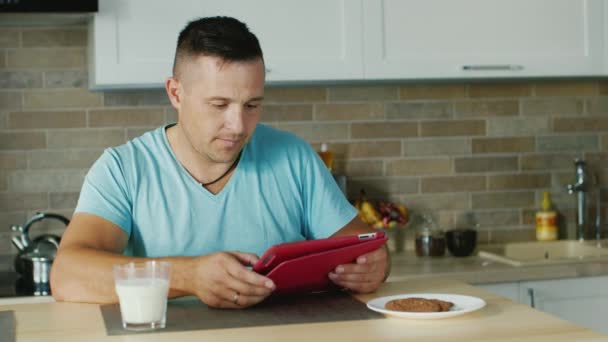 Un joven usa una tableta en la cocina, de pie junto a un vaso de leche. Estilo de vida saludable — Vídeo de stock