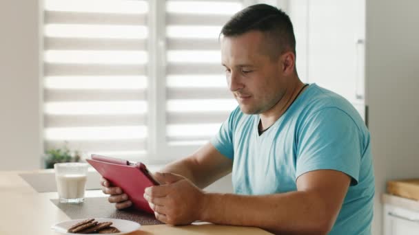 年轻人阅读数字平板电脑上的新闻。坐在厨房的窗边 — 图库视频影像