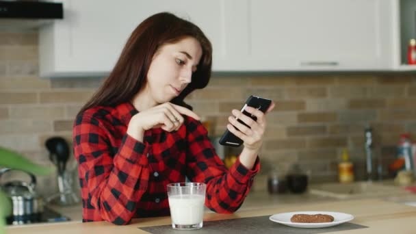 Una mujer joven usa un teléfono inteligente. Está sentado en su cocina, de pie junto a un vaso de leche. Estilo de vida saludable — Vídeo de stock