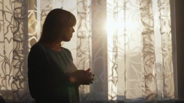 Genç kadın evde çay ya da kahve içiyor. Gün batımında pencerede dikiliyor. Evin konforu ve sıcaklığı