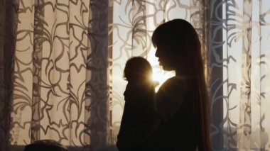 pencerede bir çocuk ile genç bir annenin silueti. Bebeği kollarına alıyor. Gün batımında