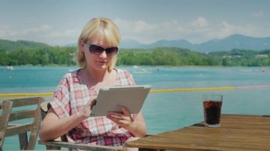 Tatilde teknoloji. Göl ve dağların arka plan üzerinde bir kafede bir masada oturan tablet ile kadın. İspanya'da tatil köyü