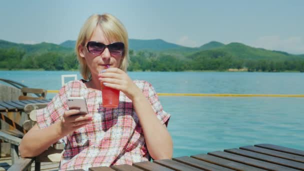 女人在咖啡店的一个夏天休息.它使用的电话。在背景上,湖水和群山.西班牙的度假胜地 — 图库视频影像