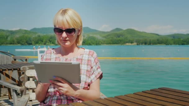 Mladá žena s tabulkou. Sedí v kavárně na terase na pozadí malebného jezera a zelených hor. Španělsko — Stock video