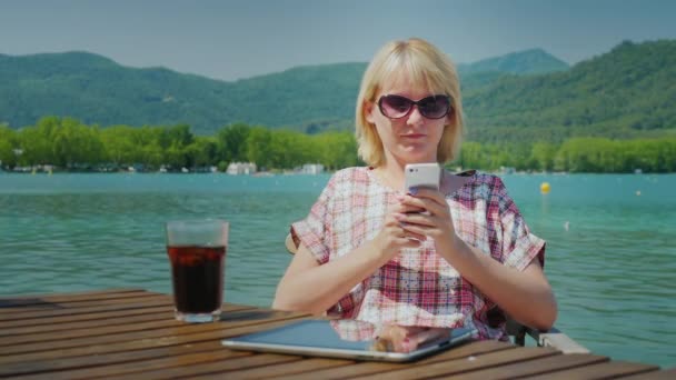 Молода туристична жінка відпочиває в прекрасному місці біля озера та гір. Він використовує телефон, сидячи за столом у кафе — стокове відео