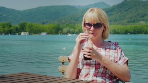 Young tourist woman relaxing in a beautiful location by the lake and mountains in Spain. Drinks Coke from a glass with a straw — Αρχείο Βίντεο