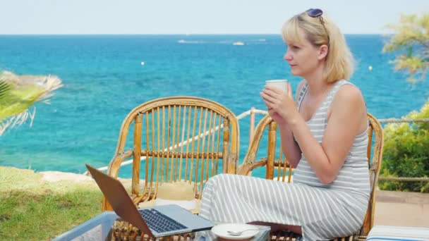 젊은 여자가 커피를 마십니다. 테라스에 앉아 바다를 내려다 보며 노트북 옆에 앉아 있습니다. 완벽 한 프리랜서 — 비디오