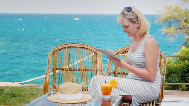 Νεαρή γυναίκα απολαμβάνει την ταμπλέτα. Καθισμένος στην καλοκαιρινή βεράντα καφέ με θέα στη θάλασσα — Αρχείο Βίντεο