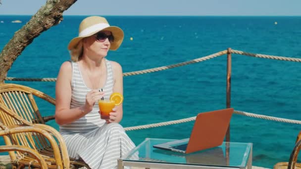 Kobieta turysta relaksujący się w kawiarni z widokiem na morze i picia koktajlu. W pobliżu jest laptop na stole — Wideo stockowe