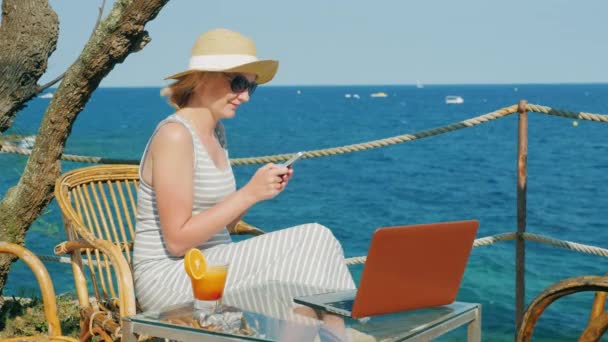 Turista mujer relajándose en un café con vistas al mar, disfruta de un teléfono inteligente, de pie junto a un ordenador portátil — Vídeo de stock