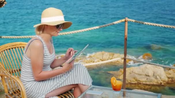 Επιχείρηση γυναίκα στις διακοπές, κάθεται σε ένα καφέ με γραφική θέα στη θάλασσα. Λειτουργεί με tablet — Αρχείο Βίντεο