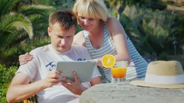 Una coppia sposata con un tablet obschaetsya hanno un video, sorridendo alla telecamera. Sullo sfondo di palme in un resort tropicale — Video Stock