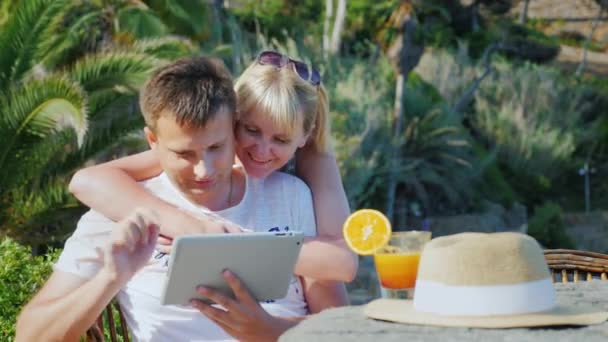 Pareja joven hablando con una tableta, video chat. Contra el telón de fondo de plantas tropicales y palmeras. Siempre conectado en vacaciones — Vídeo de stock