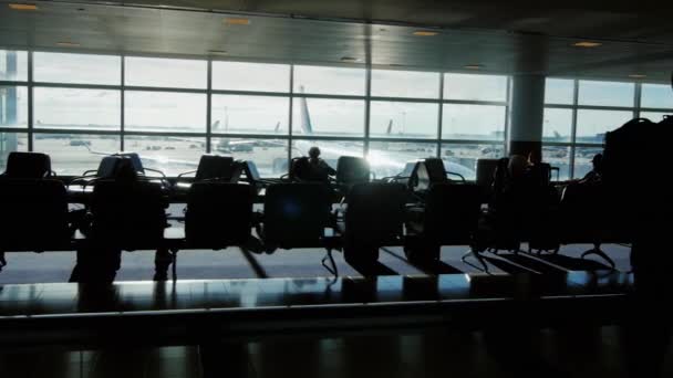 Una gran terminal del aeropuerto en un día soleado. Siluetas de personas esperando un vuelo, el avión a través de la ventana dan el hermoso resplandor del sol — Vídeo de stock